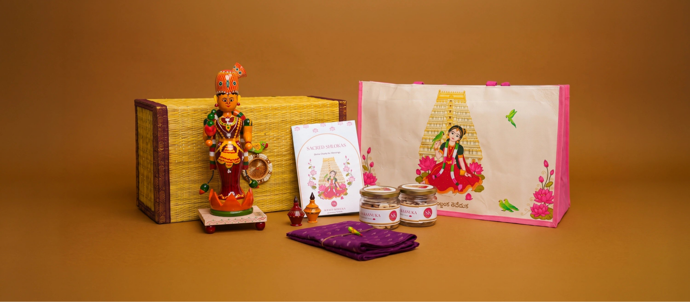 A decorative gift set arranged on a warm brown studio background, including a small colorful figurine, a woven box, a printed card, two small jars, a folded purple cloth, and a large cream tote bag with pink trim and floral artwork.